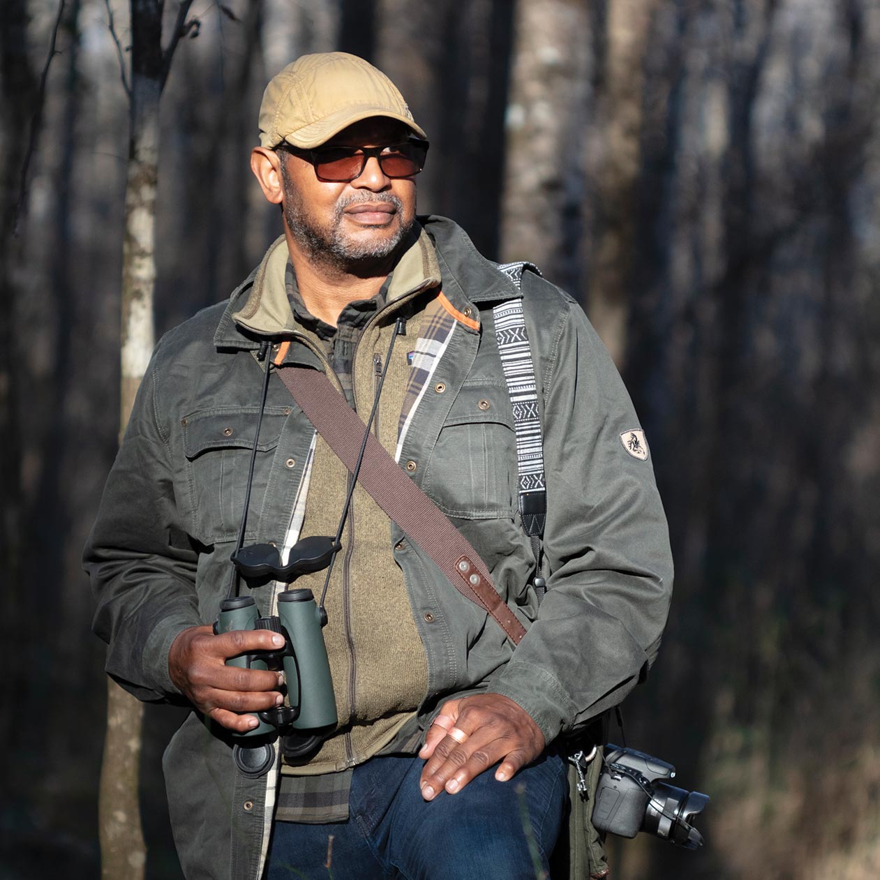 Drew Lanham (ornithologist, writer). a man holds binoculars and carries a camera in a forest setting