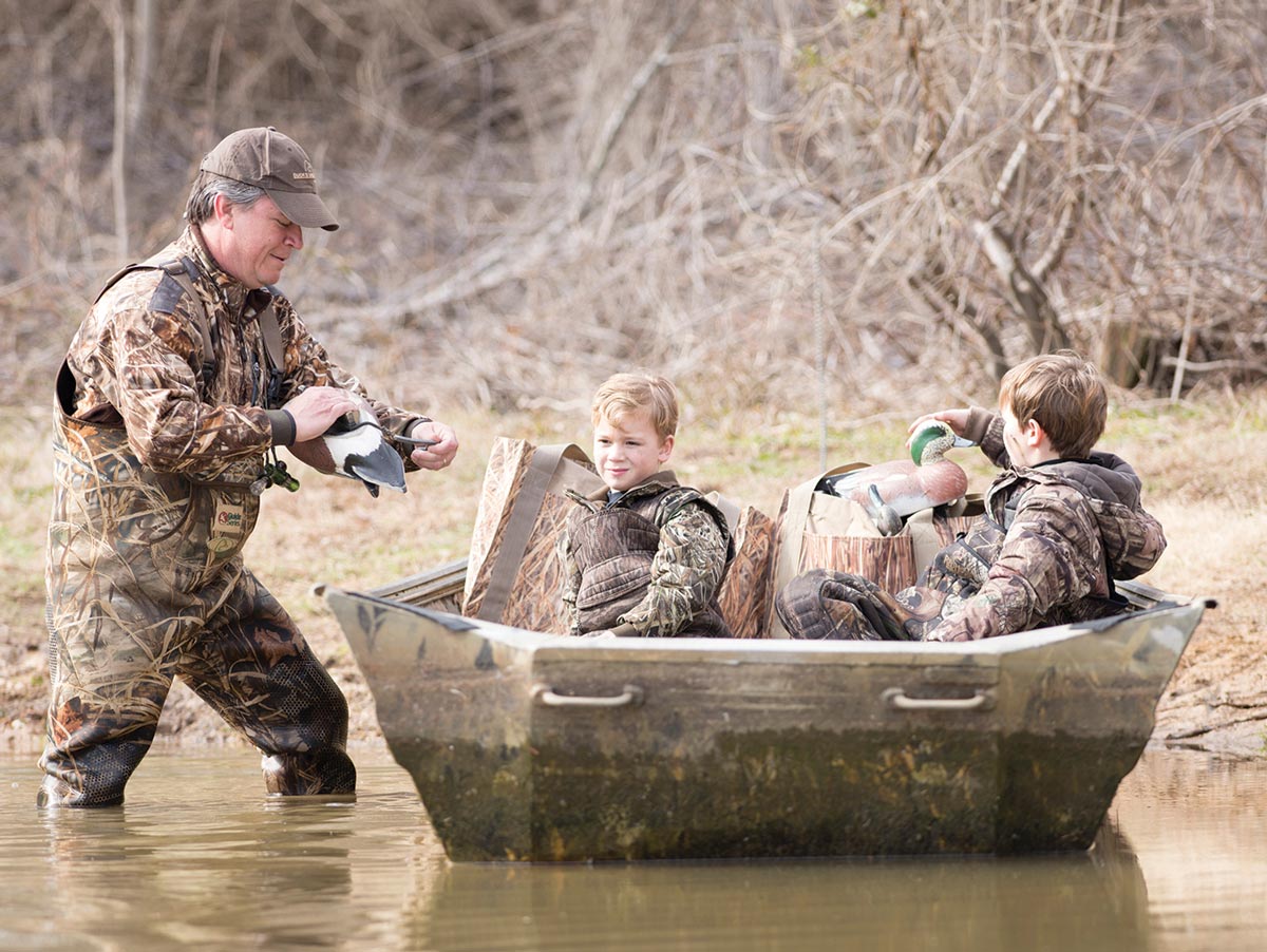 a man and two boys in camouflage load duck decoys into a flat-bottomed boat