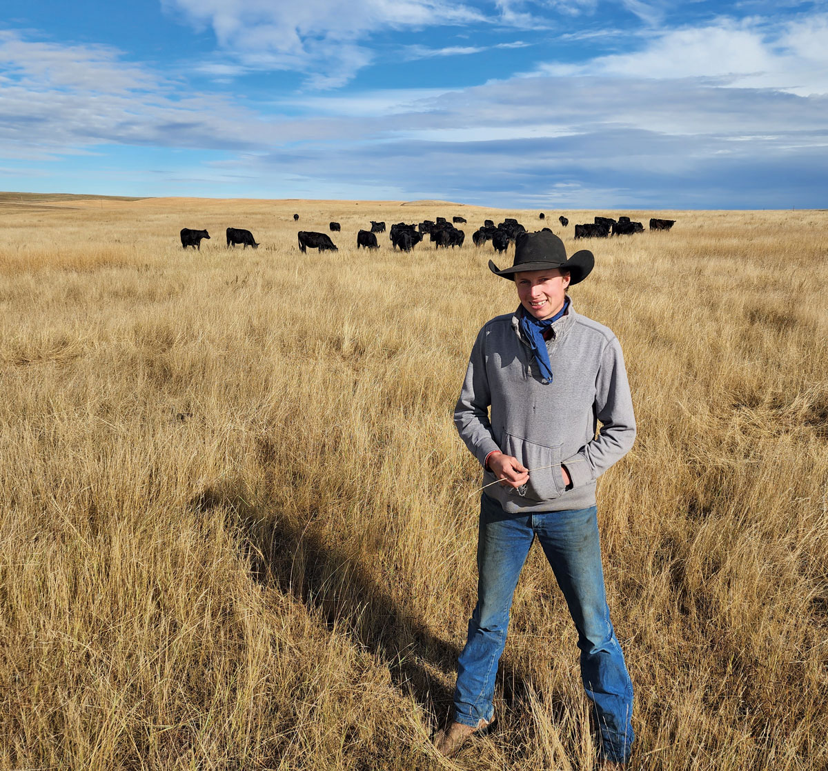A man in a cowboy hat stands in a grassy field with cows in the background.