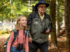 A young girl and older man on a nature walk with backpacks and binoculars around their necks.
